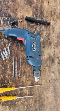 Üzerinde matkap uçları, vidalar ve tornavidalar bulunan rustik ahşap tezgah üzerine yerleştirilmiş mavi ve siyah bir elektrikli matkabın yakın görüntüsü, bir inşaat veya DIY ortamı ortaya koyuyor..
