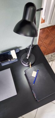 Siyah lambalı minimalist siyah bir masa, kapalı bir laptop, yansımalı dijital bir saat, bir defter, bir kalem ve yapışkan notlar, hepsi düzgünce masa yüzeyine dizilmiş..