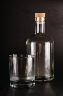 İçki ya da alkol için tasarlanmış temiz, boş bir 750 ml cam şişe, uygun bir cam bardağın yanında duruyor. Her ikisi de desenli siyah bir zemine karşı konumlanmış. Şişenin ahşap bir mantarı var..