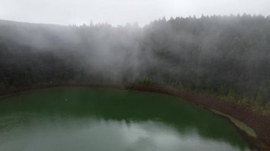Azores 'de sakin bir gölü çevreleyen sisli bir ormanın büyüleyici bir hava perdesini deneyimleyin..