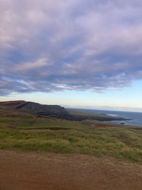 Bulutlu bir gökyüzü ufka doğru uzanır, altında mavi sularla biten çimenli bir tepe yatar.