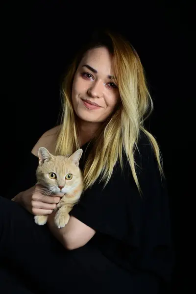 Siyah arka planda sarı bir kedi tutan sarışın bir kadının portresi.