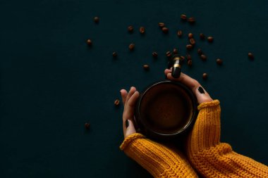 Woman's hands in a yellow knitted sweater holding a coffee mug on a dark teal background clipart