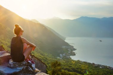 Genç bir kadın, körfez manzarasına bakan bir kayanın üzerinde huzur içinde oturuyor.