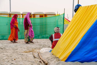 İki kadın tuvaletin önünden geçerken, bir işçi çadırın yanında oturuyor, Kumbh Mela 'nın koşuşturması ve koşuşturması arasında, kutsal bir toplantıda günlük rutinlerin zıtlığını sergiliyor..
