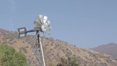 Metal bir yel değirmeni, açık mavi gökyüzünün altında yuvarlanan tepelerin zeminine karşı dimdik ayakta duruyor..