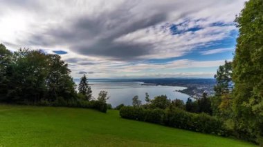 Tepenin tepesinden sakin bir gölün panoramik sonbahar manzarası, yemyeşil çimenler, canlı ağaçlar ve parçalı bulutlu bir gökyüzü..