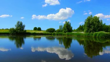 Durgun bir göl içeren sakin bir yaz manzarası pofuduk bulutları ve parlak mavi gökyüzünün altındaki yemyeşil ağaçları yansıtıyor..