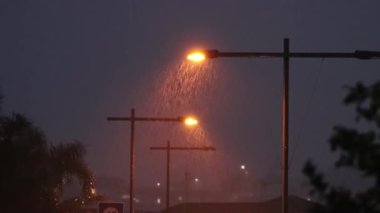 Gece vakti, sokak lambalarıyla aydınlatılmış bir şehirde yağan yağmur görüntüsü.
