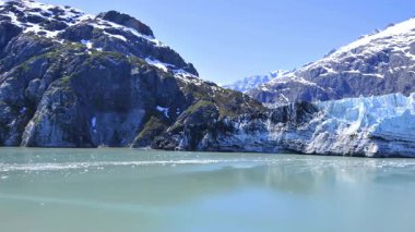 Alaska buzulunun karla kaplı dağlarla çevrili sakin, turkuaz sulara dönüşümünün çarpıcı panoramik görüntüsü..
