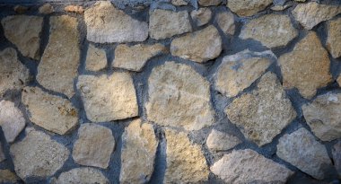 Roughly shaped stones blend harmoniously in a textured wall illuminated by warm afternoon rays. clipart