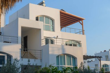 Geniş pencereli, bembeyaz bir villa yeşil bitkiler ve açık gökyüzüyle çevrili altın saatin tadını çıkarıyor..