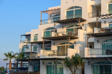 Şık bir bina, okyanus manzaralı, palmiye ağaçları ve açık mavi gökyüzü çerçeveli balkonlar içerir..