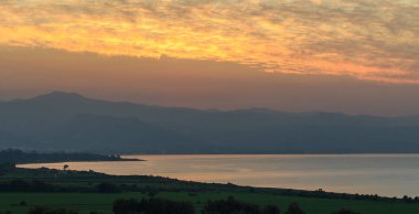 Altın renkler, yumuşak bulutlarla harmanlanır. Güneş, dingin bir göl üzerinde yuvarlanan tepeler arasında batar..
