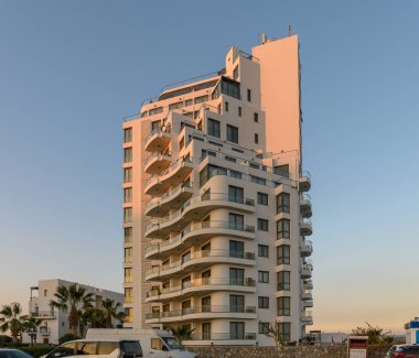 Benzersiz balkonları olan çağdaş bina, canlı bir sahil ortamında gün batımının altın renklerini yakalar..