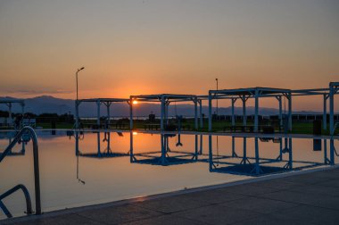 Sakin sular, canlı günbatımını yansıtıyor. Siluetler havuz kenarında rahatlarken, huzurlu bir akşam ortamı yaratıyor..