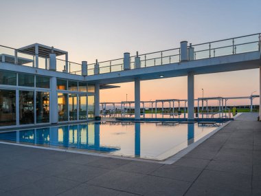 Su, günbatımının ılık tonlarını yansıtıyor. Konuklar deniz kenarındaki modern bir havuz başında dinlenirken..