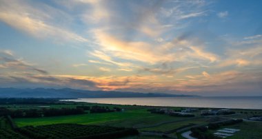 Günbatımı yemyeşil tarlaların ve sakin suların üzerine sıcak bir ışıltı saçar, uzak dağlar ve renkli gökyüzü ile yan yana gelir..