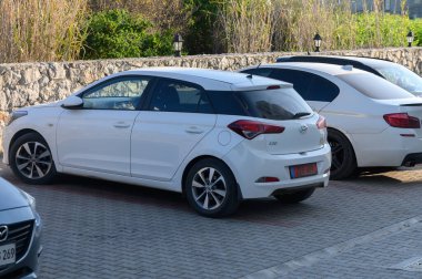 Gaziveren Kıbrıs Rum Kesimi 03.02.2025 Taş duvarlar ve yeşillik yakınlarındaki parlak güneş ışığı altında gümüş bir sedanın yanına park etmiş parlak beyaz bir hatchback..