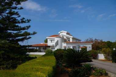 Göz kamaştırıcı beyaz bir villa, canlı yeşillik ve açık mavi gökyüzü ile çevrili, öğleden sonra zarif bir şekilde duruyor..