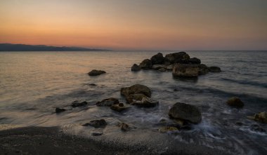 Dalgalar günbatımının yumuşak renklerini yansıtan huzurlu bir alacakaranlık gökyüzünün altında kayalık kıyılarda nazikçe tur atar..