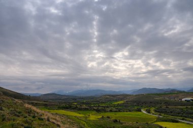 Bereketli yeşil alanlar ve dolambaçlı yollar bulutlu gökyüzü ve sakin bir manzaradaki uzak tepelerle tezat oluşturuyor..