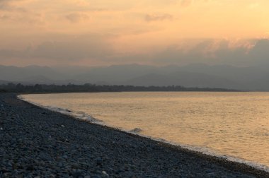 Sakin sular günbatımının ılık tonlarını yansıtır. Sahil zarif bir gökyüzü altında kıvrılır..