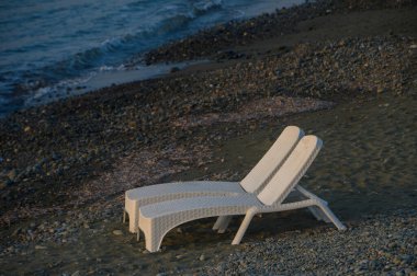 İki beyaz şezlong huzur içinde kıyıda otururlar, alacakaranlıkta hafif dalgalara bakarlar, sakin bir yansıma ve sakinlik çağırırlar..