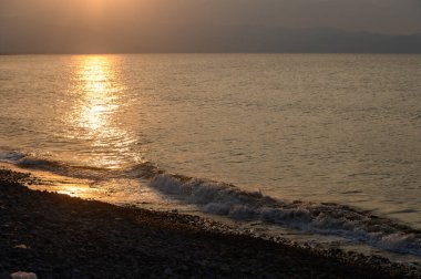 Altın ışınlar hafif dalgalar üzerinde dans eder çakıllı bir plaja yuvarlanırken alacakaranlık derinleşirken ve gündüz geceye geçerken.