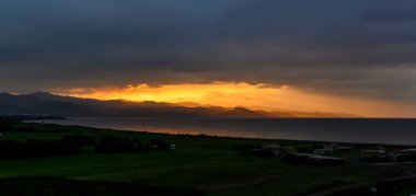 Sıcak altın renkler ufku aydınlatıyor bulutlar sakin denizin ve yeşil kıyı şeridinin üzerinde toplanırken.