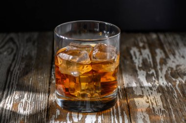 Bir bardak viski, içi buz dolu, yıpranmış ahşap bir yüzeyde oturur ve akşamın sıcacık parıltısını yakalar..