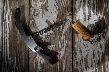 Şık bir tirbuşon, şarap mantarının yanında, yukarıdan gelen yumuşak doğal ışıkla aydınlatılmış, yıllanmış ahşap kalasların üzerinde yatar..