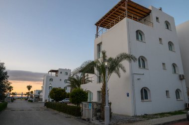 Parlak beyaz binalar kıyıda durgun durur. Avuçlar güneş batarken usulca sallanır. Huzurlu bir şekilde parıldar..