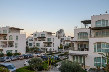 Akşam ışığı gökyüzünü yumuşak renklerle boyarken, sakin bir sahilin yanında şık beyaz daireler yükseliyor..