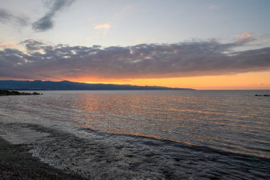 Güzel bir günbatımı, sakin denizin üzerinde canlı renkler bırakır. Uzak dağlar gökyüzüne karşı siluet oluşturur..