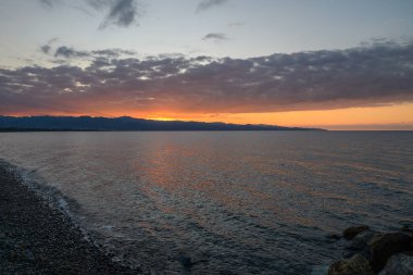 Güneş uzak dağların ardında batarken dalgalar yavaşça koyu çakıl taşlarına çarpar ve gökyüzünü ılık renklerle boyarlar..