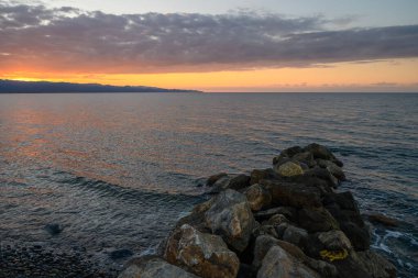 Altın güneş ışığı alacakaranlıkta hafif dalgalardan yansıyarak engebeli kıyı şeridi boyunca sakin bir atmosfer yaratır..