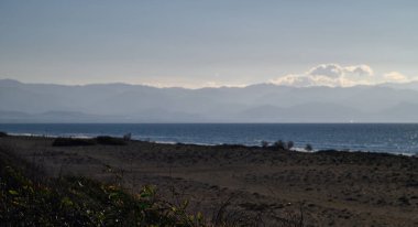 Alacakaranlıkta sakin bir kumsal manzarası, sakin dalgalar ve sisli dağlar uzaklarda kayboluyor..