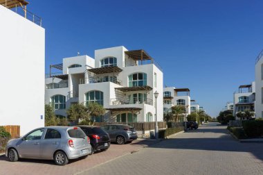 Modern beyaz binalar berrak mavi gökyüzünün altında huzurlu bir sahil havası yaratarak sakin bir yol çiziyor..