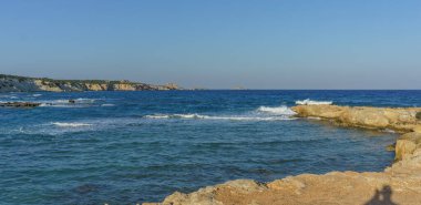 Sakin bir kıyı manzarası, alacakaranlıkta güneşli kayalara karşı parlak mavi suları ve yumuşak dalgaları barındırır..