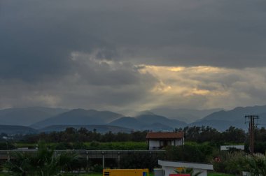 Clouds gather ominously as twilight descends, casting a soft glow over the distant mountains and peaceful homes. clipart