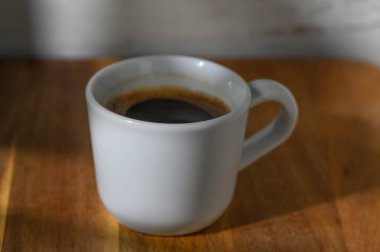 Ilık güneş ışığı, parlak ahşap bir yüzey üzerinde dinlenen küçük bir fincan koyu kahveyi yıkar ve rahatlatıcı bir anı davet eder..