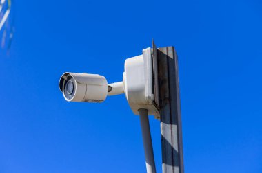 Bir güvenlik kamerası, parlak mavi gökyüzünün altındaki manzarayı gözetleyen bir direğin üzerinde tetikte bekliyor..