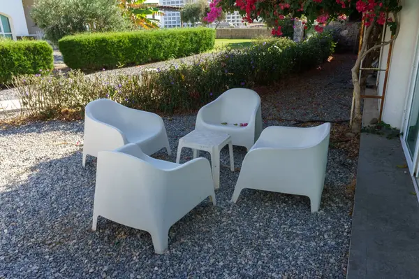 Küçük bir masanın etrafına yerleştirilmiş dört zarif beyaz sandalye, yemyeşil yaprakların ve renkli çiçeklerin arasına yerleştirilmiş..