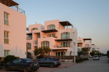 Modern beyaz binalar sakin bir cadde boyunca uzanır, akşam ışığını karşılar ve huzurlu bir ortam yaratır..