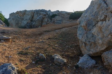 Bir yürüyüşçü, parlak bir güneşin altında, manzaralı bir yamaçta, kayalık zeminde yol alır..