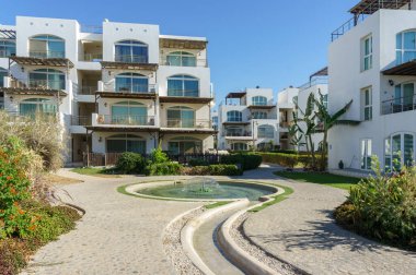 Huzurlu bir avluda, modern konutlarla çevrili, sıcak güneş ışığıyla yıkanmış bir çeşme bulunur..