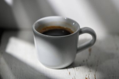 Sıcak bir kahve fincanı güneş ışığını yakalar, içerideki derin, zengin bir içeceği ortaya çıkarır, yumuşak gölgeler ve aydınlık ile çevrili..