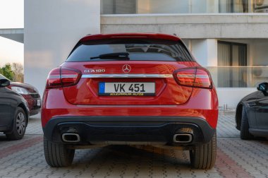 Gaziveren Kıbrıs Rum Kesimi - 30.01.2025 Altın güneş ışığı sıcak bir parıltı saçarken çağdaş binaların yakınında şık kırmızı bir SUV duruyor.