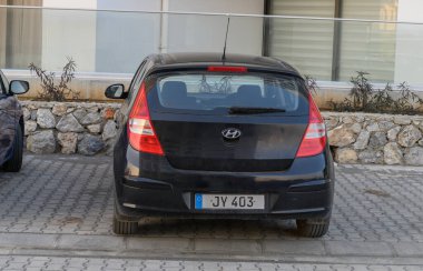 Gaziveren Kıbrıs Rum Kesimi - 30.01.2025 Siyah bir hatchback taştan bir duvara yaslanmış, sıcak öğle güneşinin altında güneşlenerek sakin bir atmosfer yaratıyor.